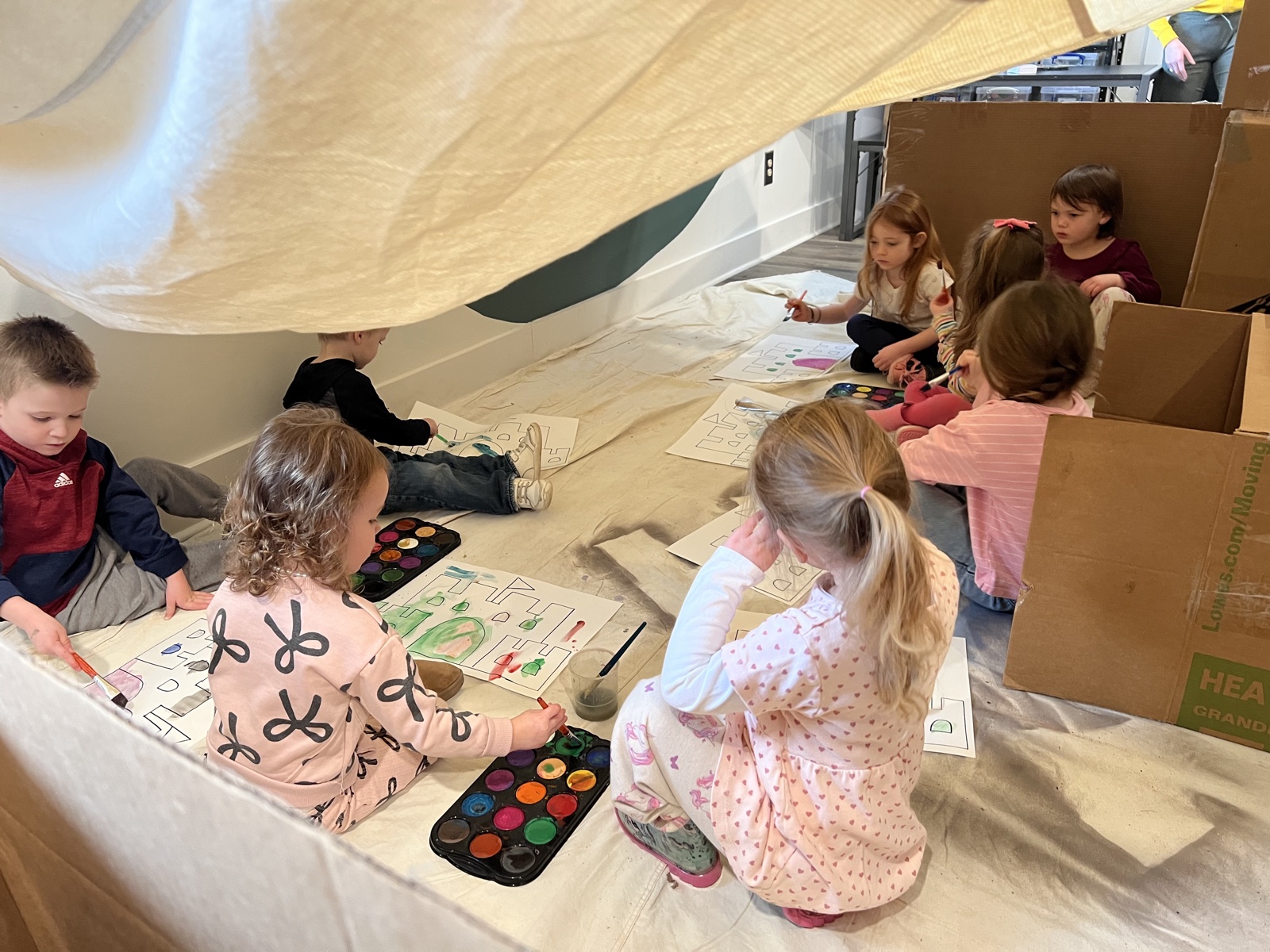 Children painting and drawing together inside a handmade cardboard fort